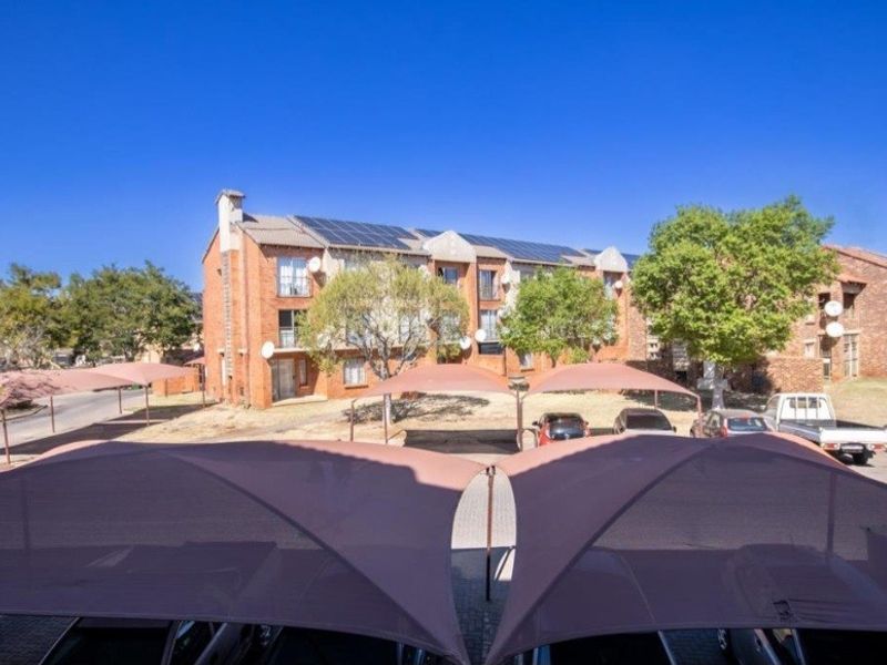 Exterior view of a multi-story brick apartment complex featuring solar panels on the roof and covered parking bays with shade cloth structures.