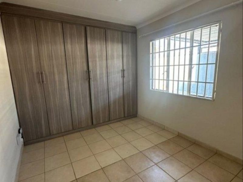 A clean, empty bedroom featuring neutral tiled flooring, built-in wooden cupboards, and a window fitted with security bars.