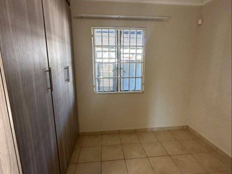 A clean, empty bedroom featuring neutral tiled flooring, built-in cupboards, and a window fitted with security bars.