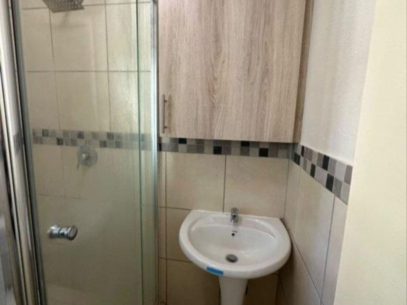 Compact bathroom featuring a pedestal basin, a glass-enclosed shower with a rain shower head, and neutral wall tiling with a decorative mosaic border.