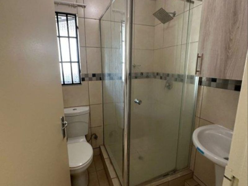 A compact bathroom featuring a glass shower enclosure, pedestal basin, toilet, and neutral wall and floor tiling.