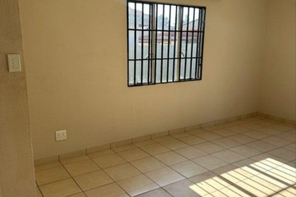 An empty living room featuring neutral tiled flooring, a window with security bars, and a curtain rail, offering a clean and versatile space.