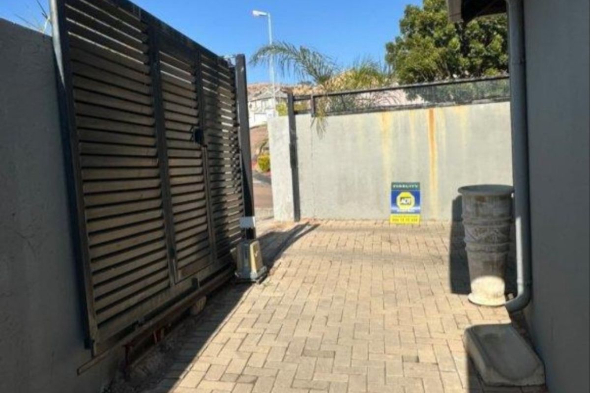A paved side access area featuring a slatted metal gate, boundary wall, and functional drainage downpipe.