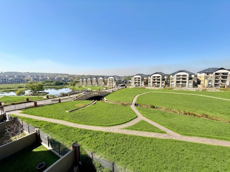 Elevated view overlooking a well-maintained residential estate featuring landscaped green spaces, paved walking paths, a stone bridge, and a scenic lake.