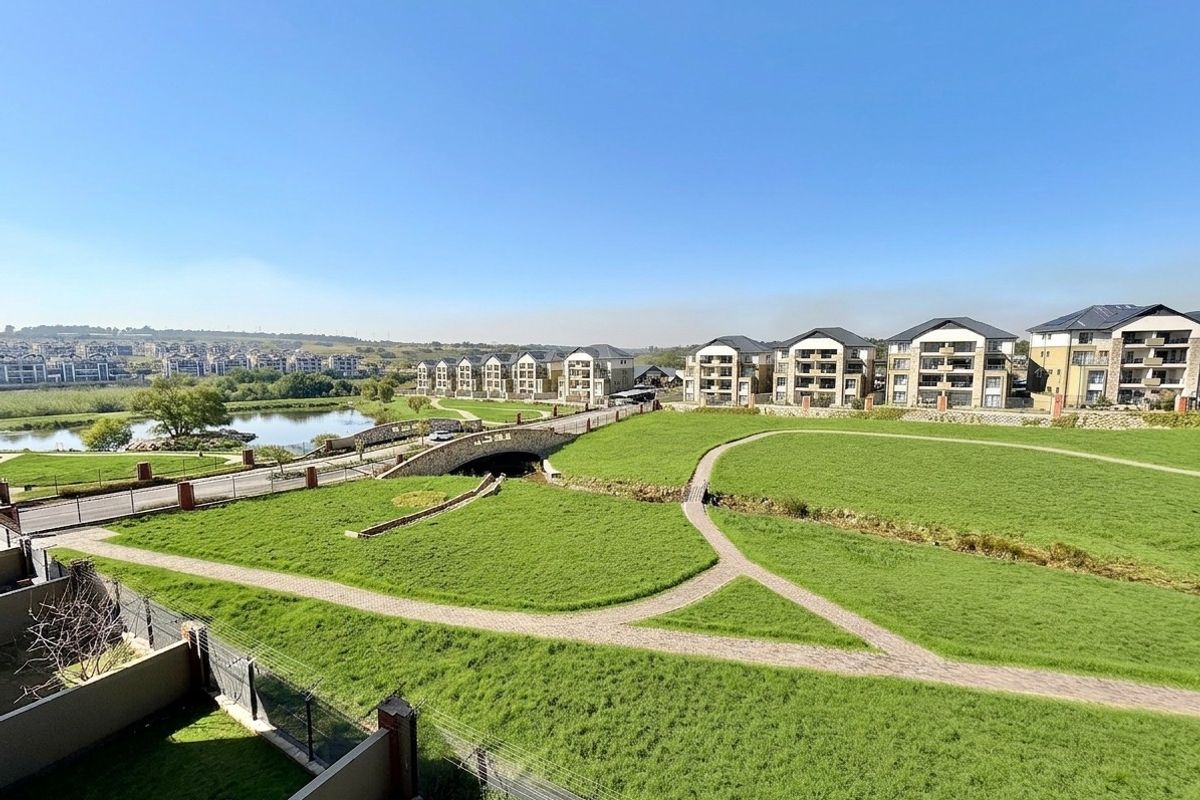 Elevated view overlooking a well-maintained residential estate featuring landscaped green spaces, paved walking paths, a stone bridge, and a scenic lake.