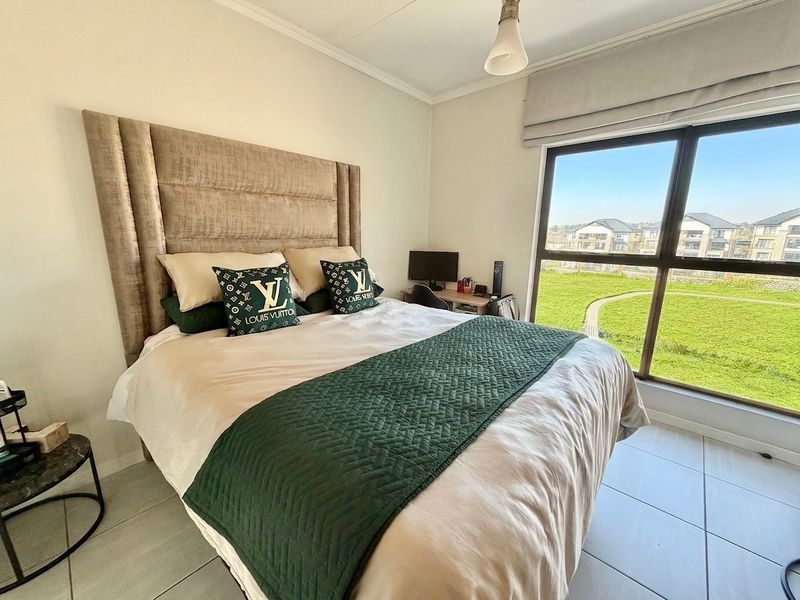 A well-lit bedroom featuring large windows with views, neutral tiled flooring, and a modern roman blind.