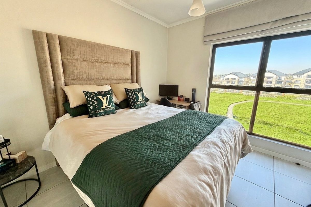 A well-lit bedroom featuring large windows with views, neutral tiled flooring, and a modern roman blind.