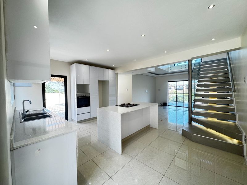 Modern open-plan kitchen featuring a central island, stone countertops, built-in appliances, and a striking staircase with glass balustrades in a bright, tiled living space.