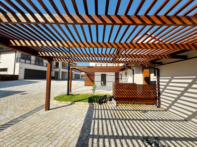 A modern outdoor area featuring a large timber pergola providing shade over a paved driveway and entrance space.