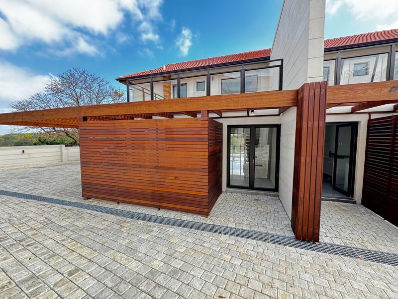 A well-maintained rear exterior featuring a timber pergola, privacy screen, paved courtyard with integrated drainage, and an upper-level balcony with glass balustrading.