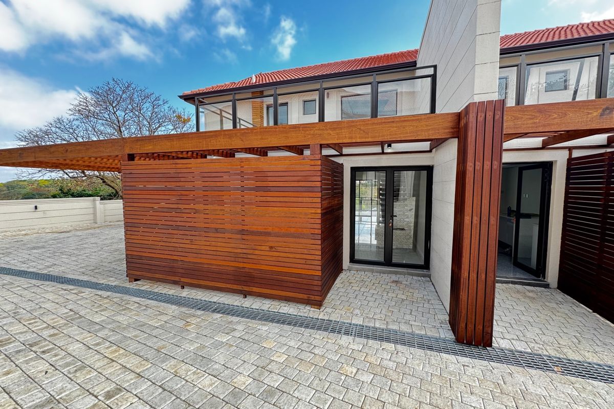 A well-maintained rear exterior featuring a timber pergola, privacy screen, paved courtyard with integrated drainage, and an upper-level balcony with glass balustrading.