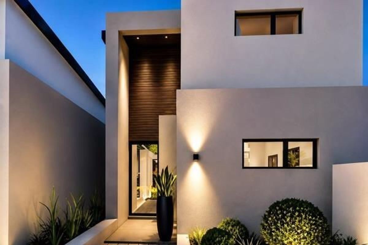 A modern, architecturally designed front facade featuring clean lines, warm accent lighting, timber cladding, and manicured landscaping.