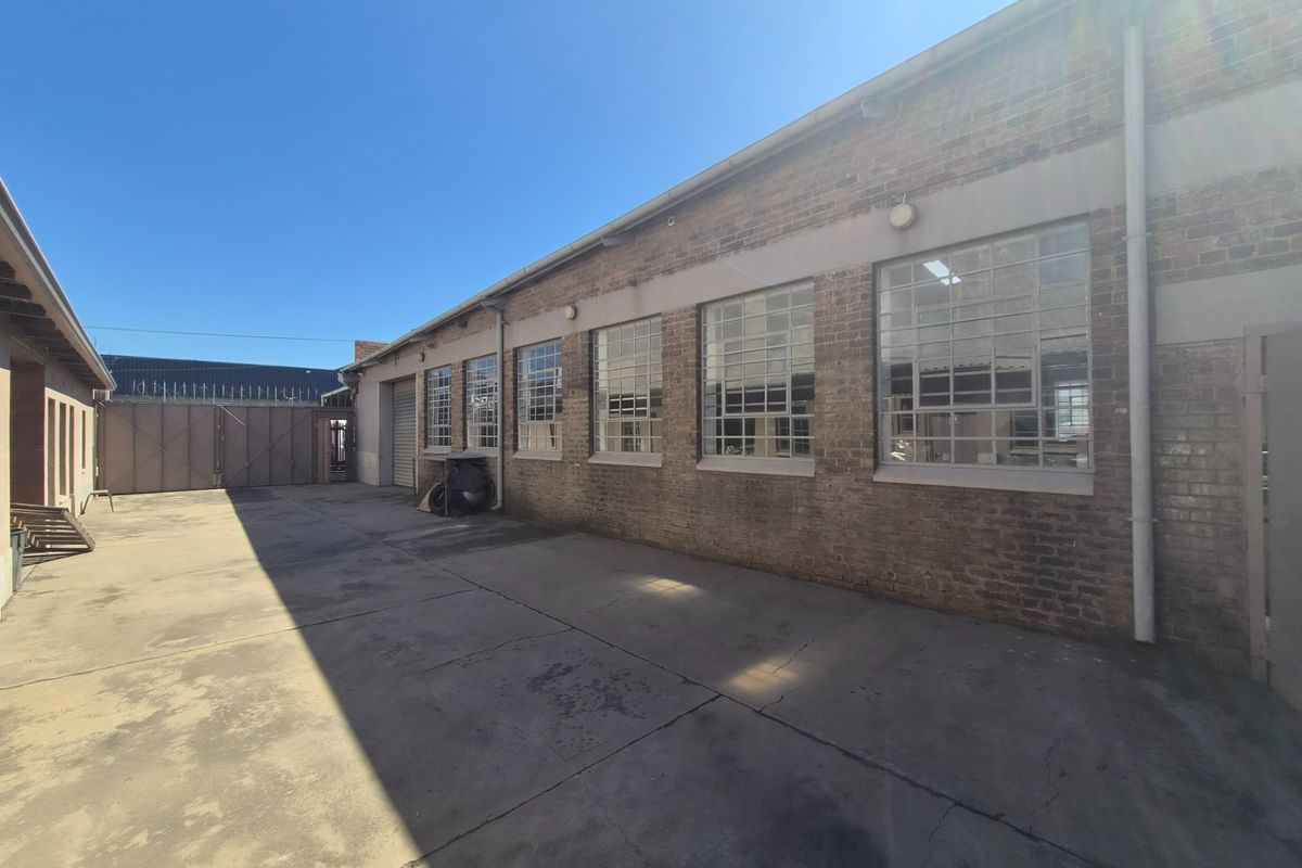 Rear industrial yard featuring exposed brick walls, large steel-framed windows with security bars, and a concrete surface with a roller shutter door access.