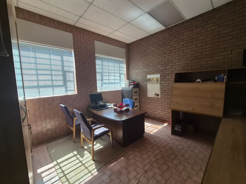 A functional office space featuring exposed brick walls, tiled flooring, and a suspended ceiling with natural light provided by two windows with venetian blinds.