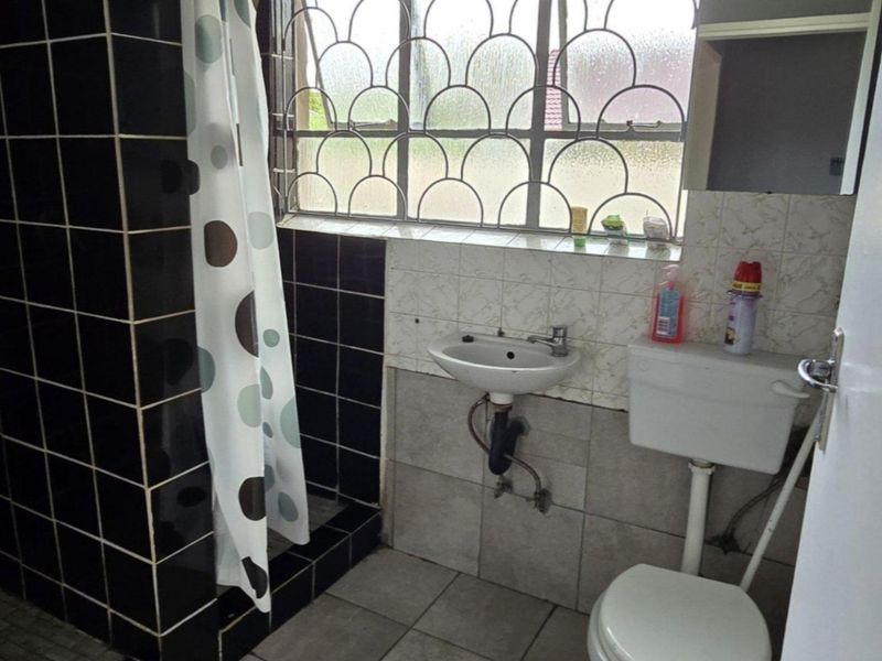 A functional bathroom featuring a shower, pedestal basin, and toilet, with tiled finishes and security bars on the window.