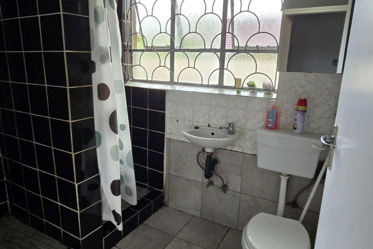 A functional bathroom featuring a shower, pedestal basin, and toilet, with tiled finishes and security bars on the window.