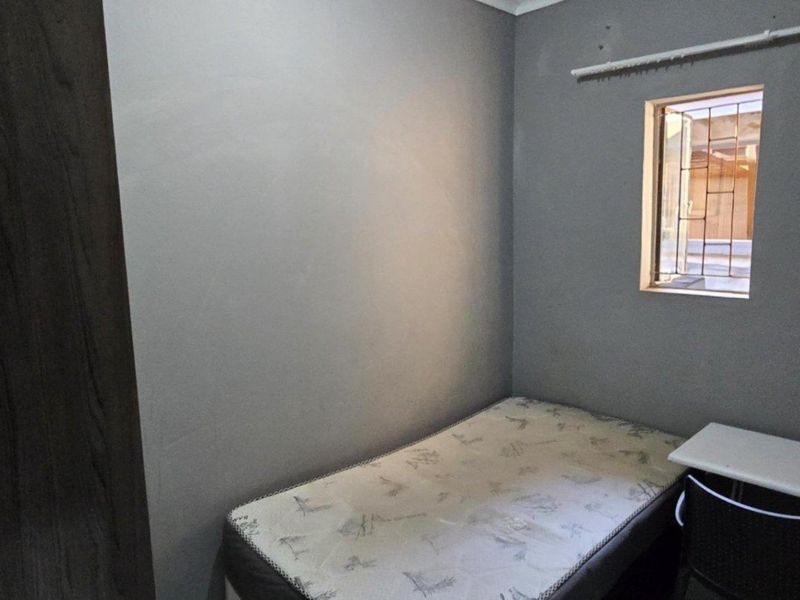 A compact bedroom featuring grey painted walls, a built-in cupboard, and a window fitted with security bars.