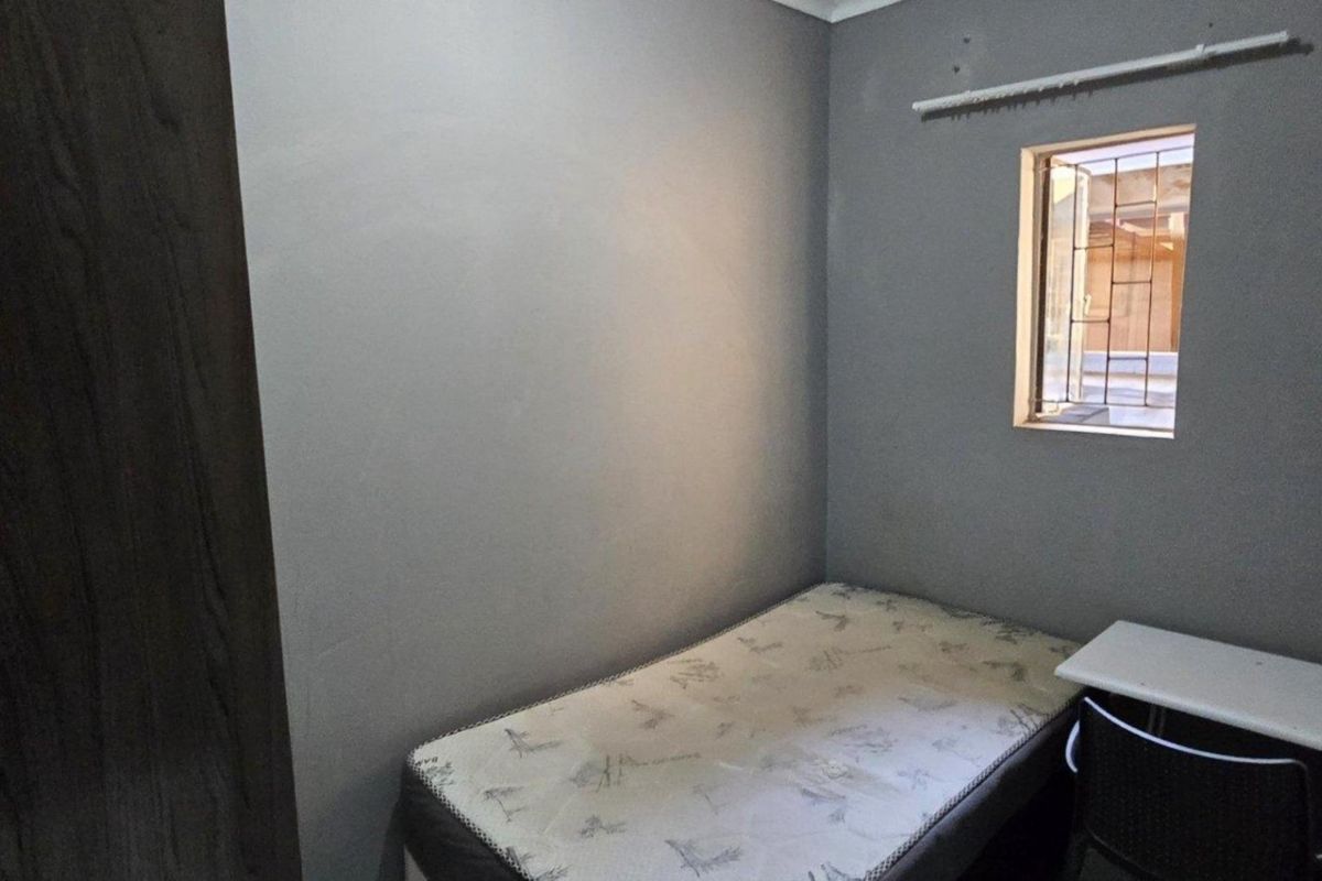 A compact bedroom featuring grey painted walls, a built-in cupboard, and a window fitted with security bars.
