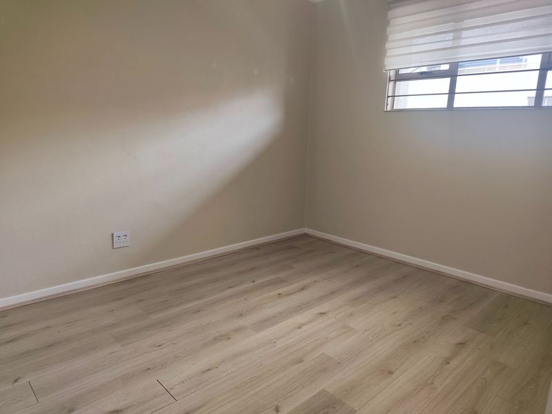 A clean, empty bedroom featuring modern light-toned laminate flooring, neutral walls, and a window fitted with security bars and a roller blind.