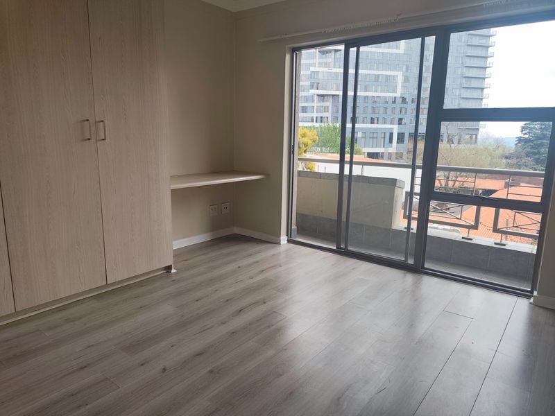 A bright, empty bedroom featuring modern laminate flooring, built-in cupboards, a fitted desk, and a sliding glass door leading to a balcony.