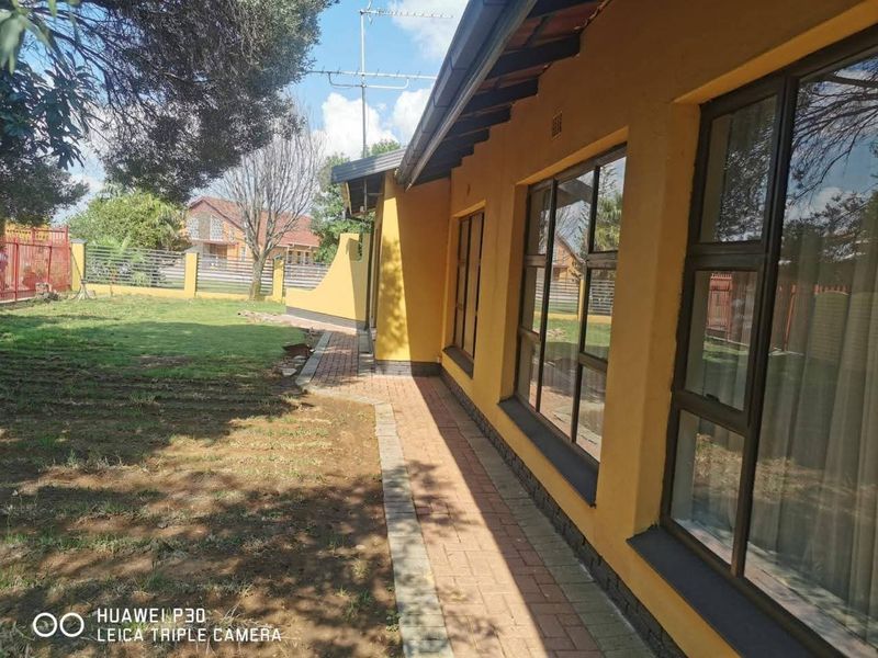 A view of the rear exterior of the house featuring a paved walkway, yellow painted facade, and aluminium window frames overlooking the garden.