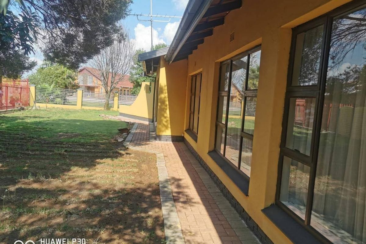 A view of the rear exterior of the house featuring a paved walkway, yellow painted facade, and aluminium window frames overlooking the garden.