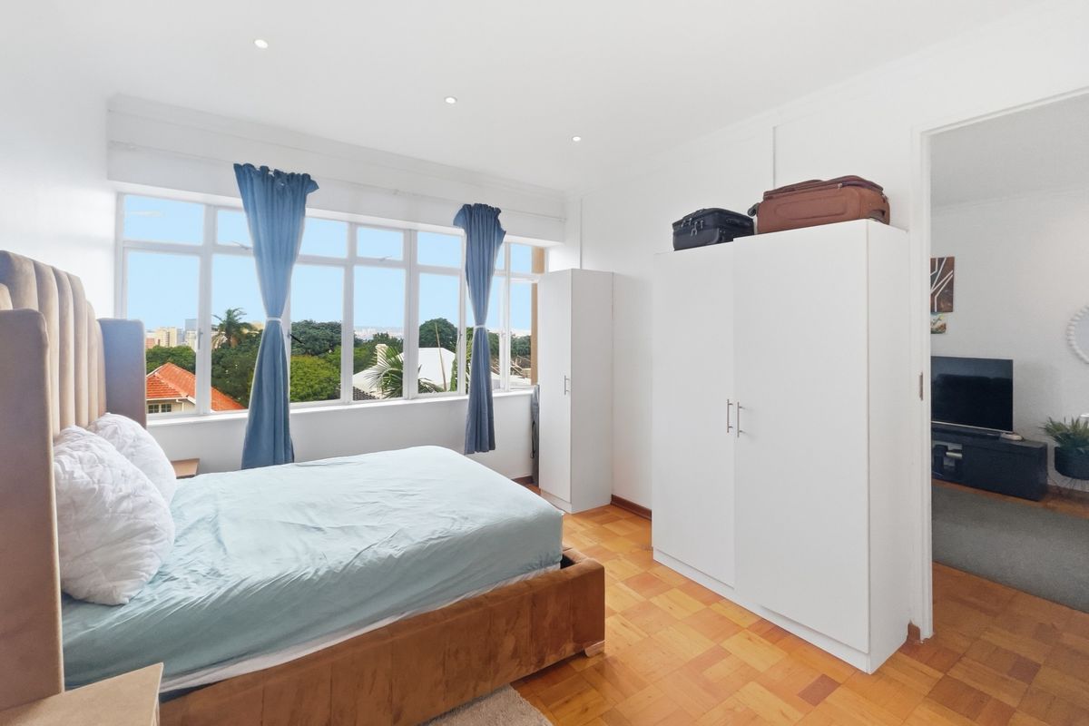 A bright bedroom featuring parquet flooring, recessed lighting, and large windows offering an elevated view of the surrounding neighborhood.