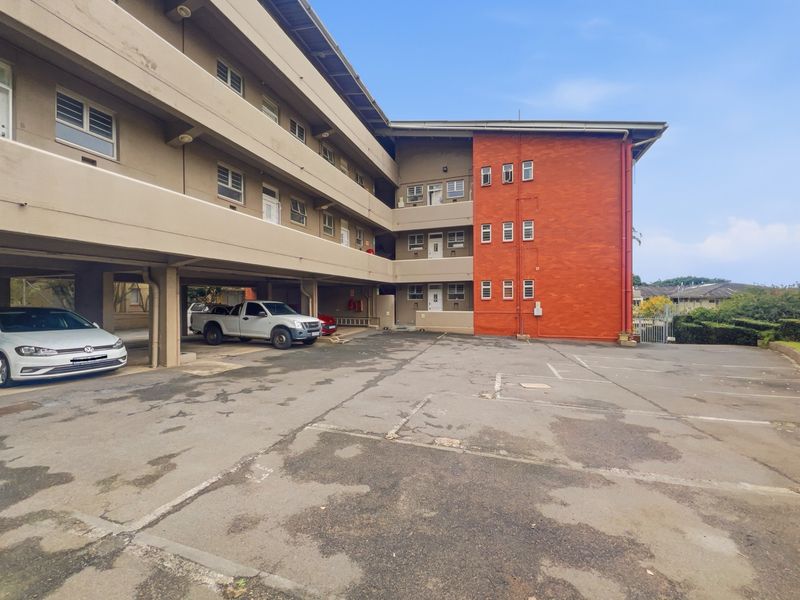 Exterior view of a multi-story apartment building featuring a paved parking area with covered bays and a prominent red brick facade.