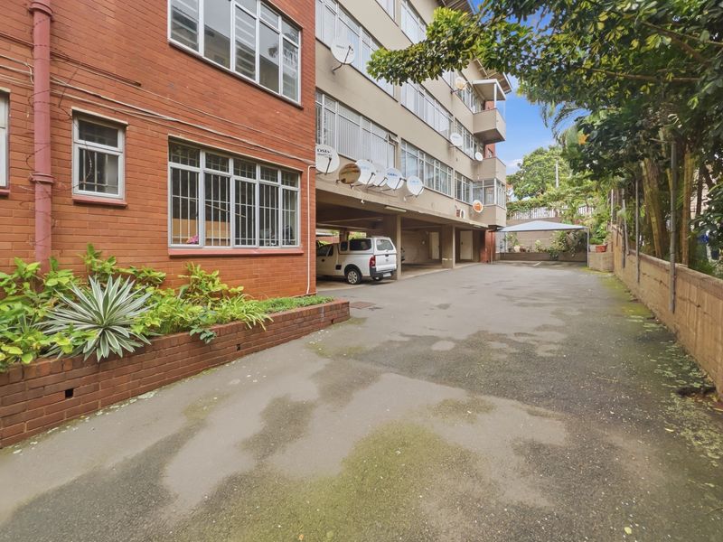 Exterior view of the apartment building featuring a brick facade, covered parking area, and a paved driveway with a brick planter box.