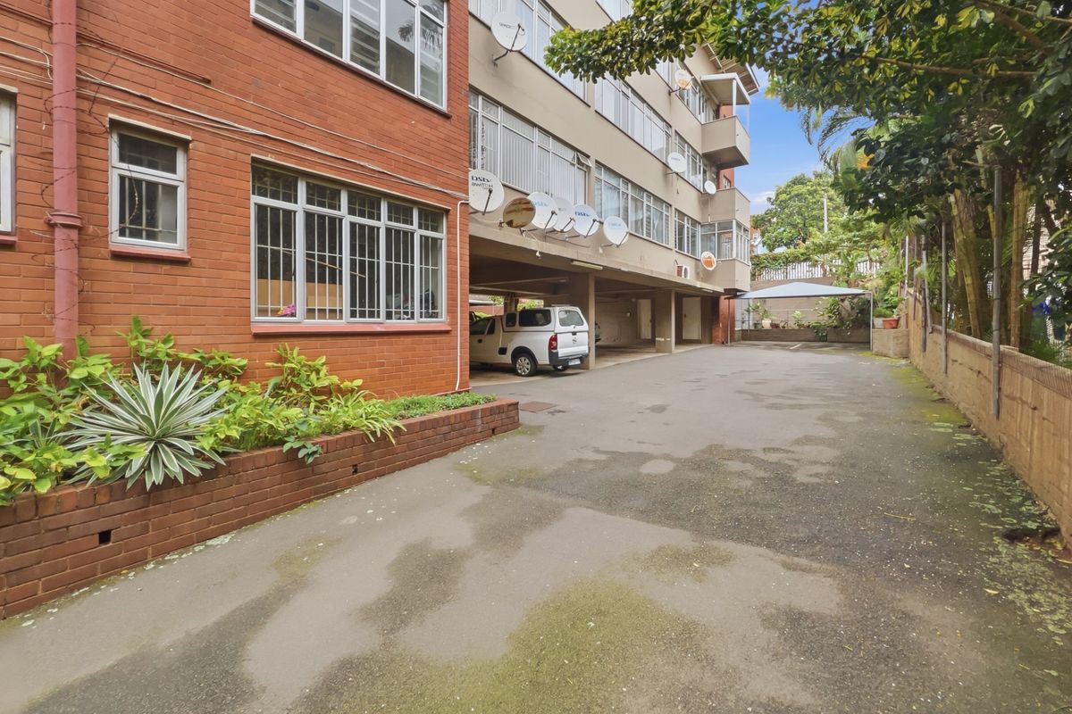 Exterior view of the apartment building featuring a brick facade, covered parking area, and a paved driveway with a brick planter box.