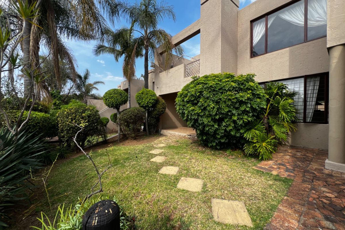 Rear view of the apartment building featuring a private garden, paved walkway, and an upper-level balcony with stucco exterior finish.