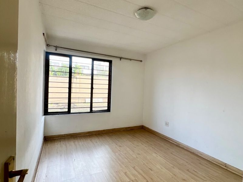 An empty bedroom featuring light-toned laminate flooring, white walls, and a window fitted with security bars.