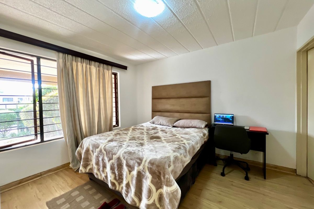 A functional bedroom featuring laminate flooring, a textured ceiling, and large windows with security bars, providing natural light and a neutral foundation.
