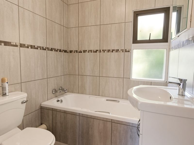 A clean, functional bathroom featuring a bathtub, toilet, and vanity basin with neutral wall tiling and a window for natural light.