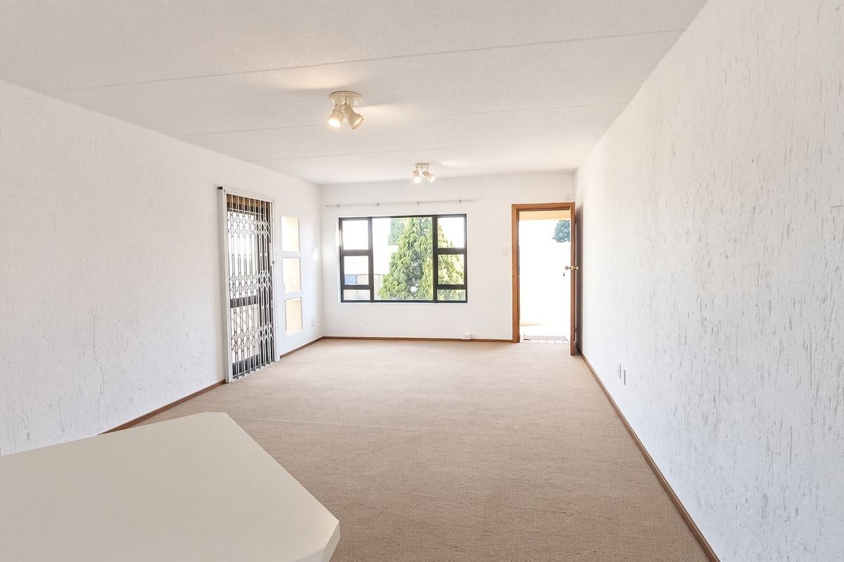 A spacious, empty living area featuring neutral carpeted flooring, textured walls, and large windows providing natural light and access to an exterior space.