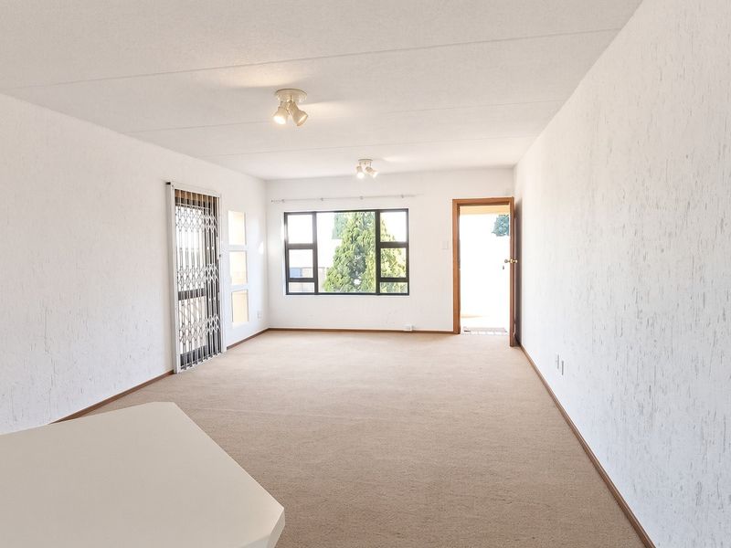 A spacious, empty living area featuring neutral carpeted flooring, textured walls, and large windows providing natural light and access to an exterior space.