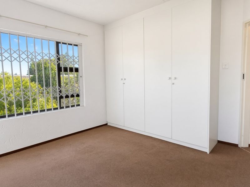 A bright, empty bedroom featuring neutral carpeted flooring, built-in cupboards, and a window fitted with a security gate.