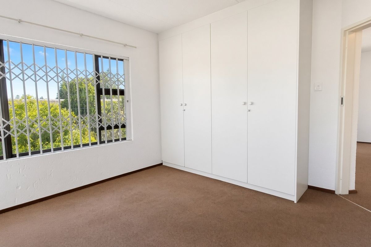 A bright, empty bedroom featuring neutral carpeted flooring, built-in cupboards, and a window fitted with a security gate.