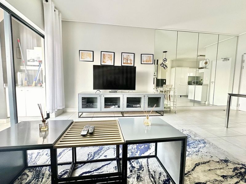 A bright, modern living room featuring large sliding glass doors, neutral tiled flooring, and a decorative mirrored wall reflecting the adjacent kitchen area.