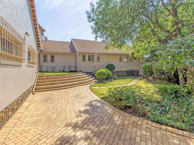 A well-maintained rear exterior view featuring a paved driveway, curved stone steps leading to the house, and a landscaped garden area.