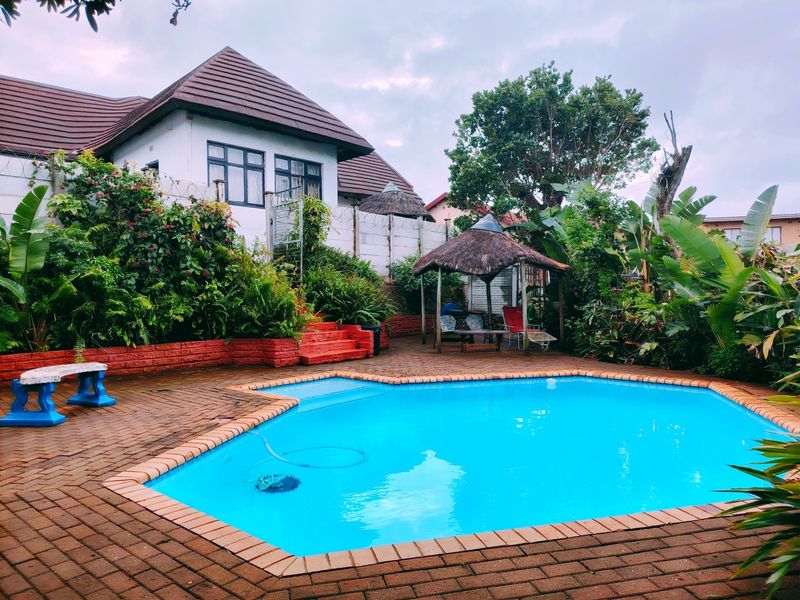 A private backyard featuring a swimming pool, brick paved surround, and a thatched gazebo set within a landscaped garden area.