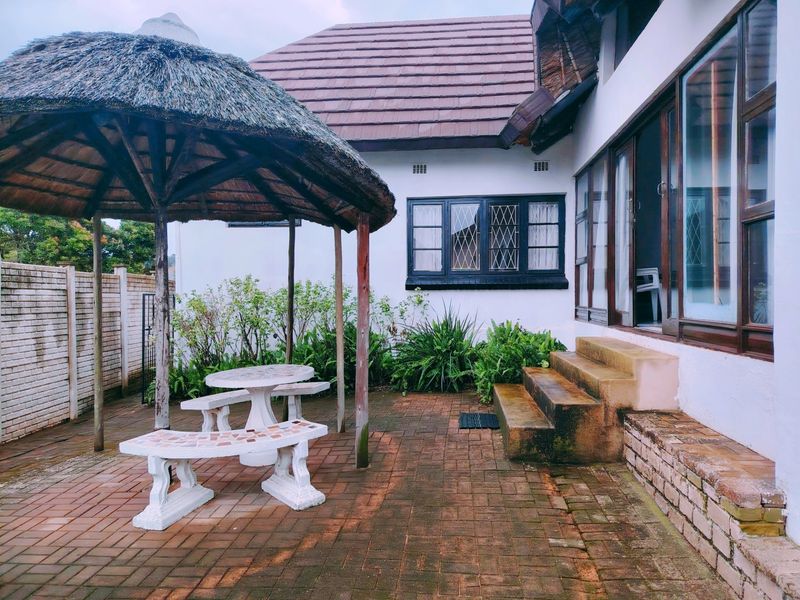 A paved outdoor patio area featuring a traditional thatched gazebo, concrete steps leading to the house, and established garden borders.