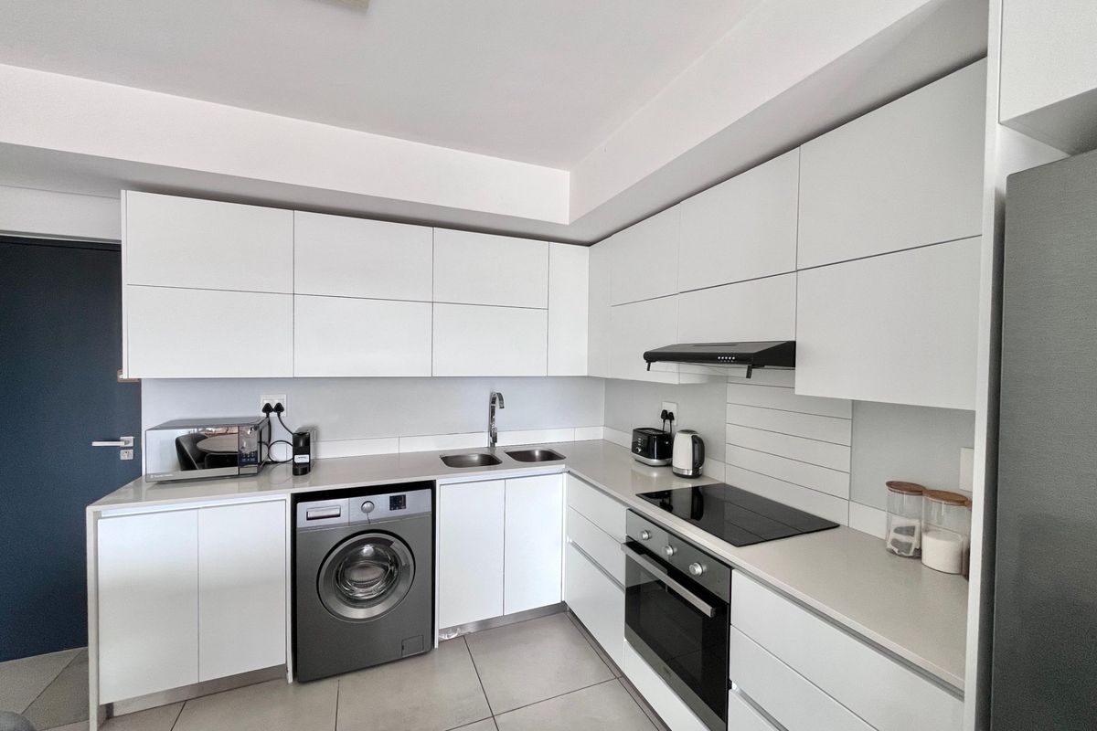 Modern kitchen featuring sleek white cabinetry, stone countertops, integrated appliances including a washing machine, electric hob, and oven with tiled flooring.