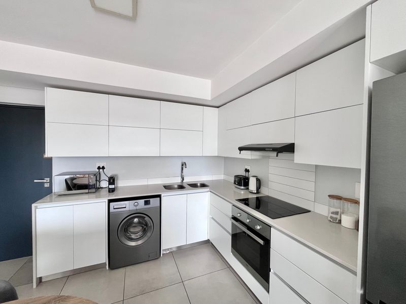 Modern kitchen featuring sleek white cabinetry, stone countertops, integrated appliances including a washing machine, electric hob, and oven with tiled flooring.