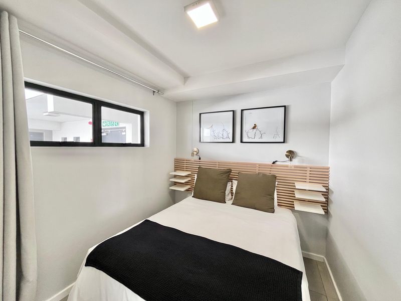 A compact, modern bedroom featuring a custom timber headboard with integrated floating shelves and a sleek black-framed window.