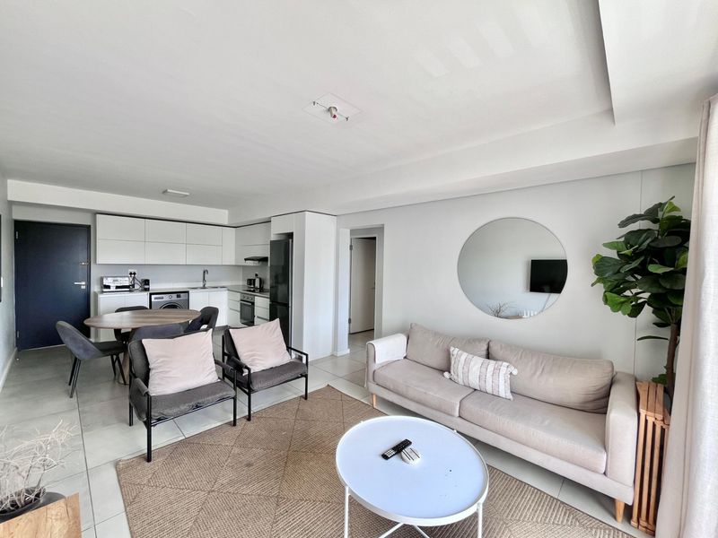 A bright open-plan living and kitchen area featuring modern white cabinetry, integrated appliances, and neutral tiled flooring throughout.
