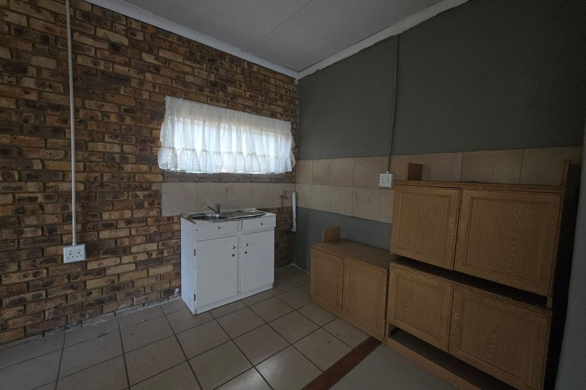 A basic kitchen area featuring exposed brick walls, tiled flooring, a single sink unit, and dated wooden cabinetry requiring modernization.