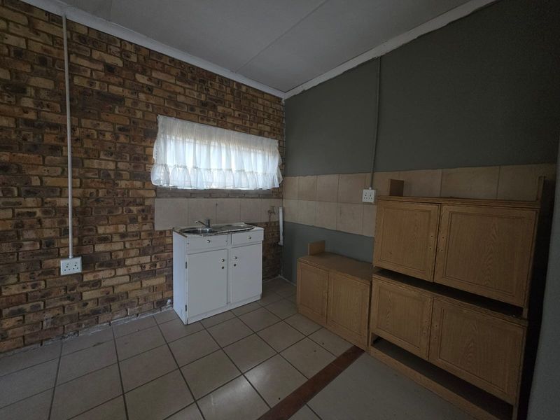 A basic kitchen area featuring exposed brick walls, tiled flooring, a single sink unit, and dated wooden cabinetry requiring modernization.
