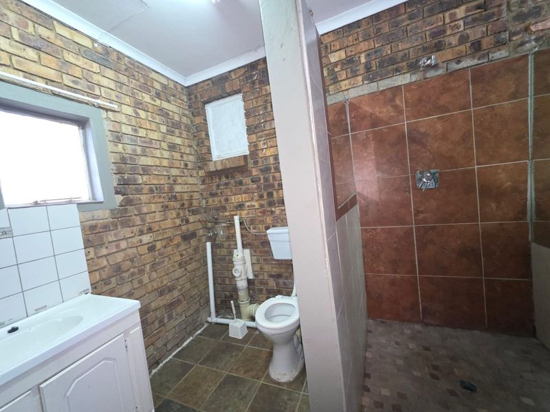 A bathroom featuring exposed brick walls, a shower enclosure, toilet, and vanity basin. The space requires renovation to update finishes and conceal exposed plumbing.