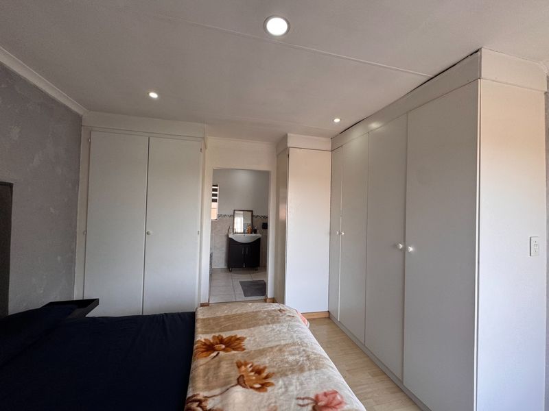 A well-maintained bedroom featuring extensive built-in cupboards, modern recessed downlighting, and laminate flooring, with an adjoining en-suite bathroom visible.